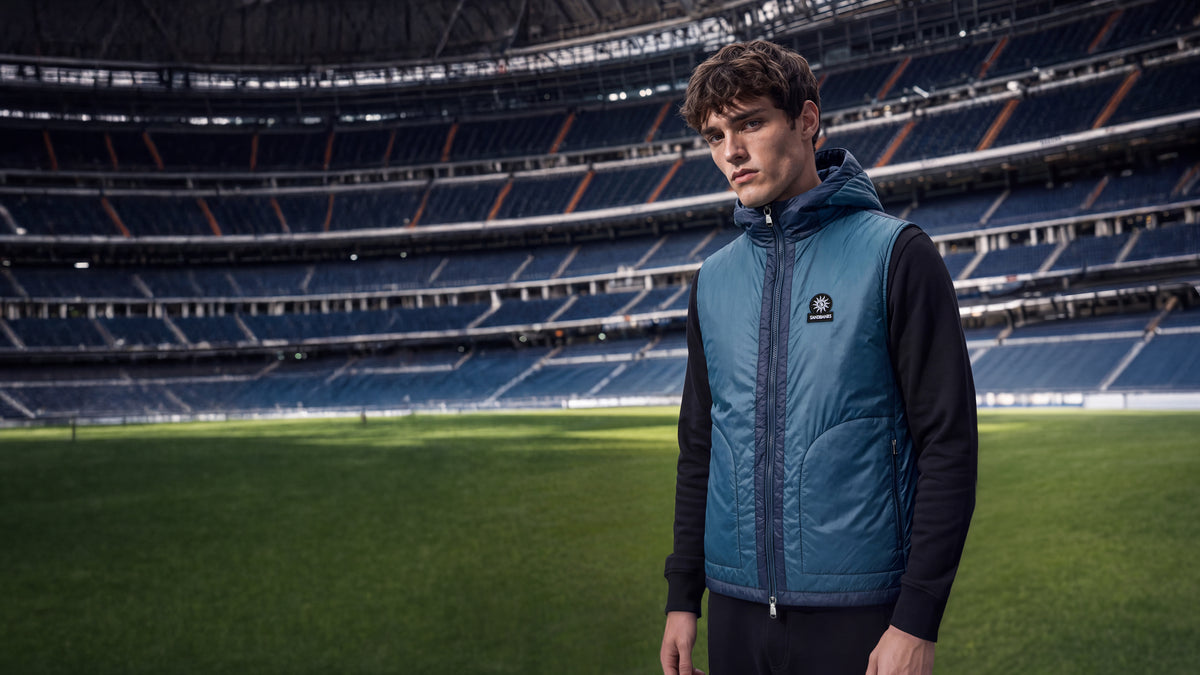 A man stands on a green soccer field inside a large, empty stadium, wearing a blue zippered puffer vest over a black long-sleeve shirt.