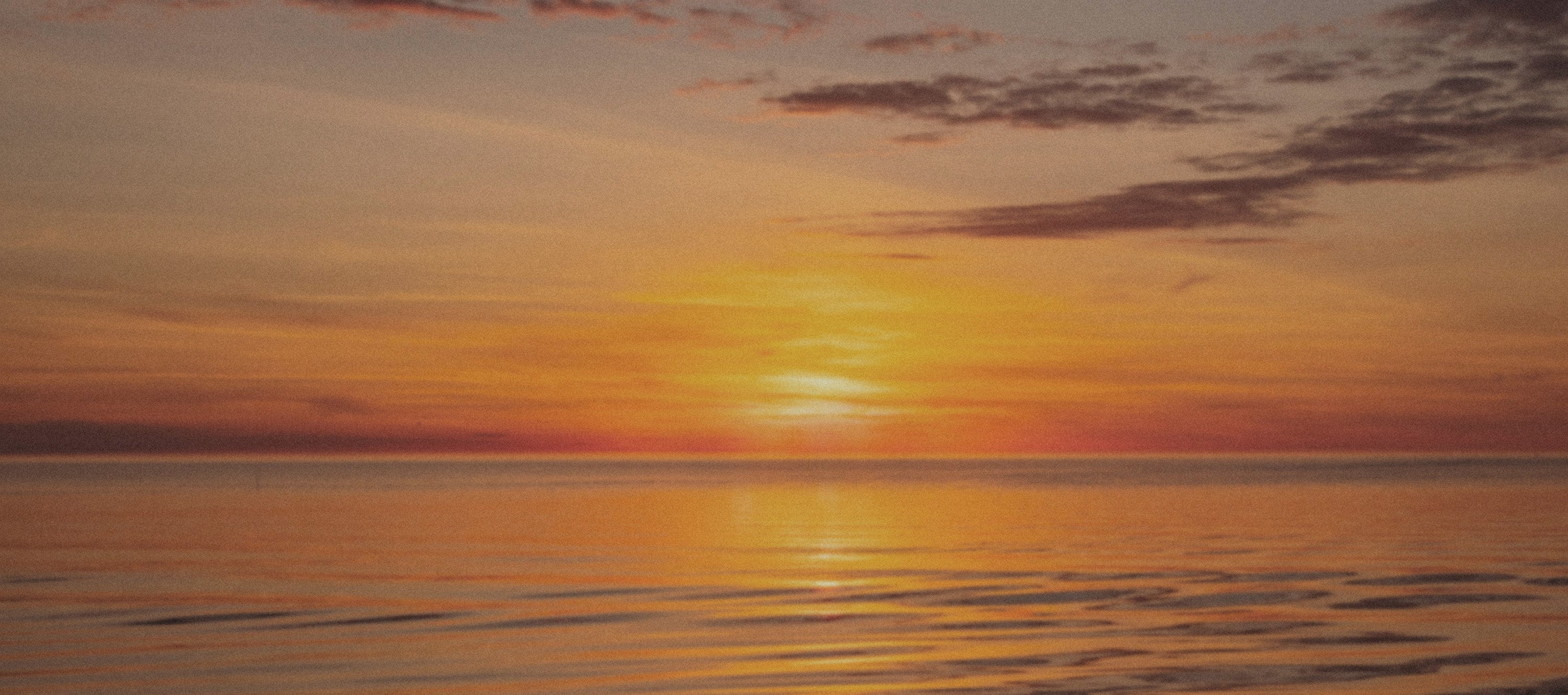 A serene sunset over calm water, with golden and orange light reflecting across the horizon and soft clouds in the sky.