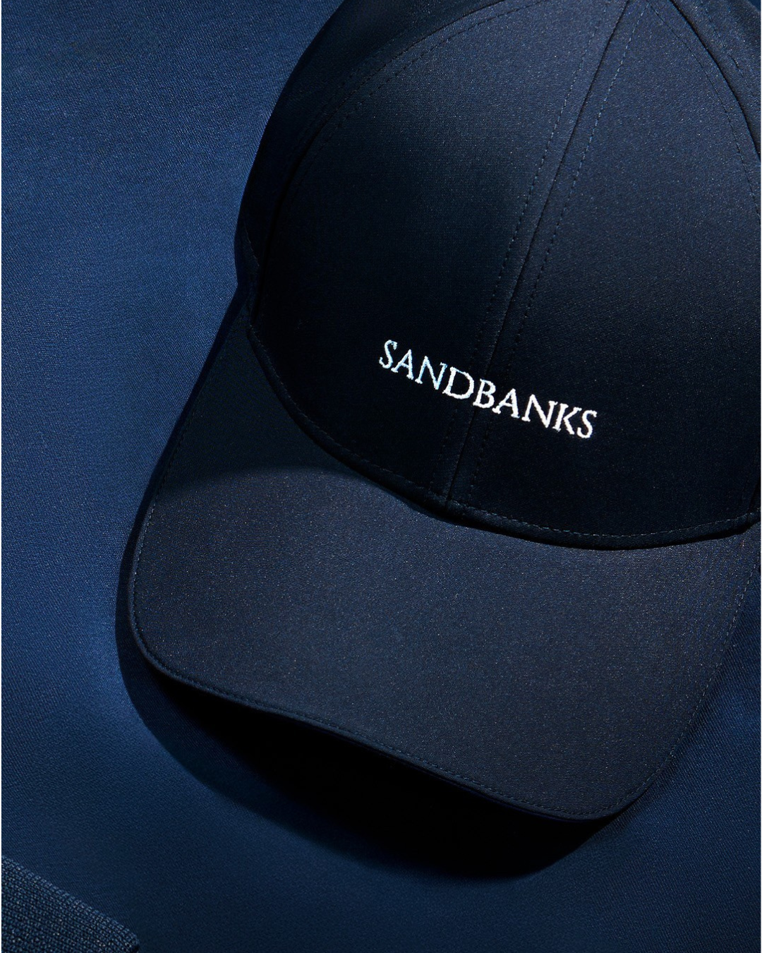 A dark navy blue baseball cap with the brand name 'SANDBANKS' embroidered in white on the front, resting on a matching navy blue textured surface.