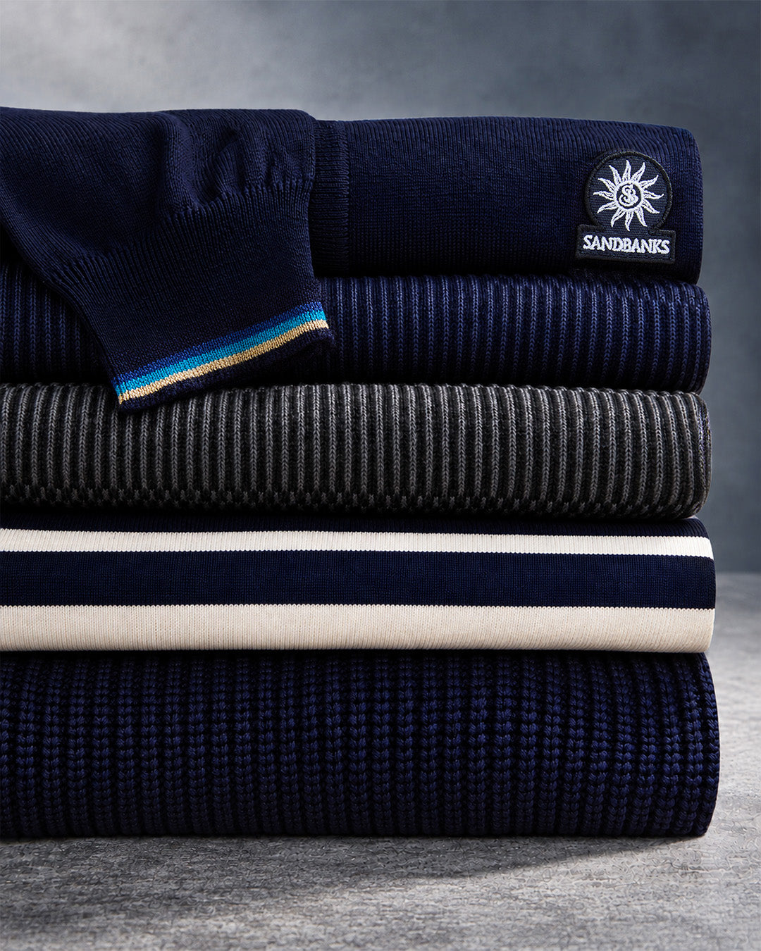 A stack of folded knit garments in multiple colors and textures, including navy blue, charcoal gray, cream, and dark blue with thin multicolor stripes. A circular SANDBANKS logo patch is visible on the top garment.