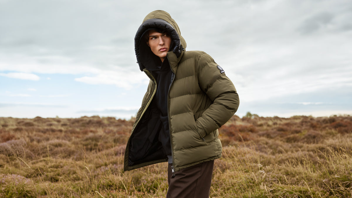 A model wearing an olive green hooded puffer jacket with a black interior lining, standing in an open grassy landscape under a cloudy sky.