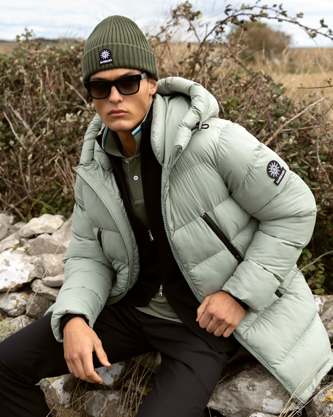 A man wearing a light sage green puffer jacket, olive green beanie, black sunglasses, and a black zip-up hoodie sits outdoors on a stone among dry grass and shrubs.