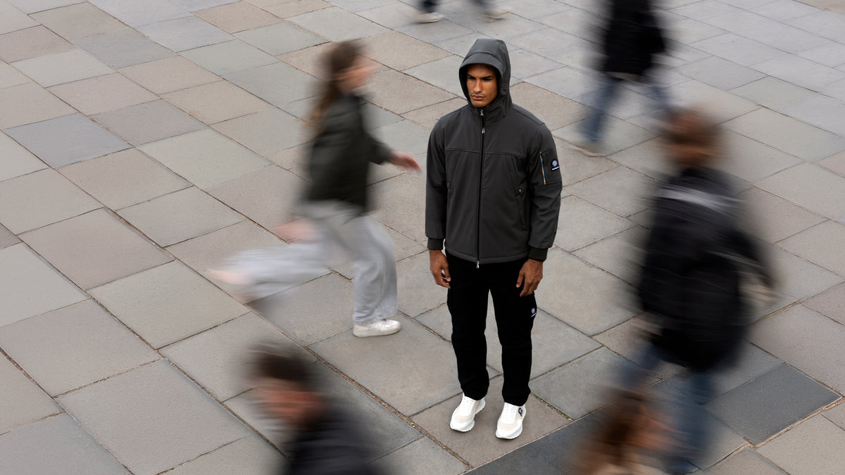 A man wearing a dark gray hooded zip-up jacket, black pants, and white sneakers stands on a paved outdoor walkway, surrounded by blurred pedestrians.