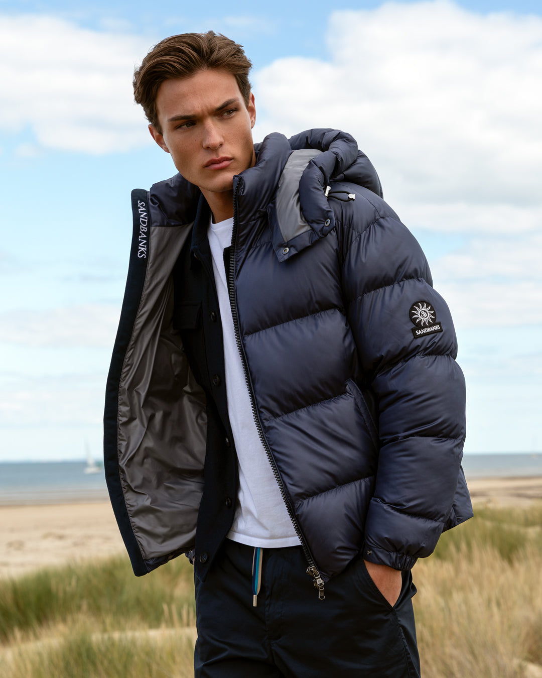 A man wearing a dark navy blue puffer jacket with a high collar and zippered front, standing outdoors in a grassy coastal area under a cloudy sky. The jacket has a visible circular logo patch on the sleeve and 'SANDNES' text on the inner collar.