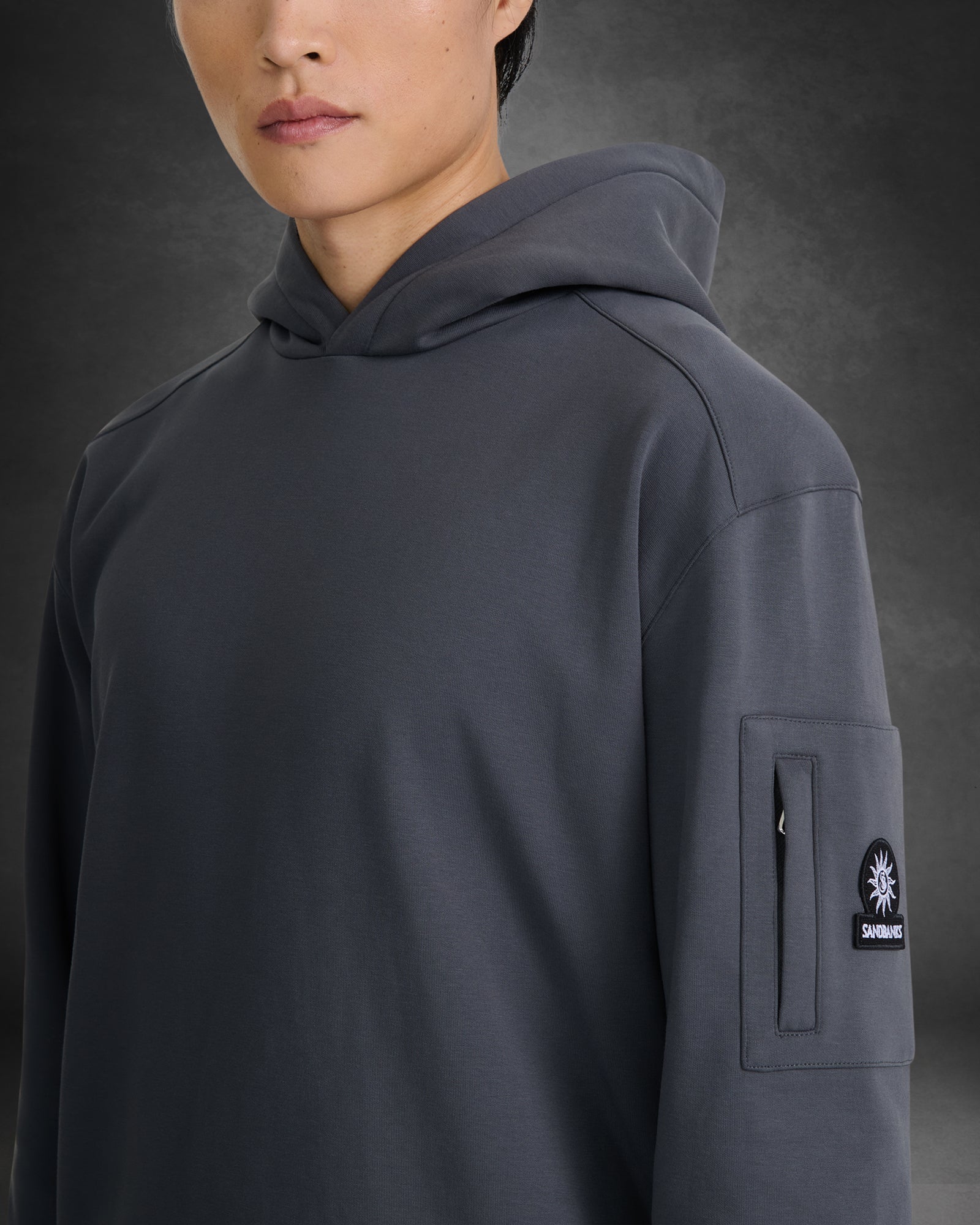 A close-up of a person wearing a dark gray hooded sweatshirt with a vertical zip pocket on the sleeve featuring a circular black patch with a white star logo and the brand name 'SKINNYBONES'.