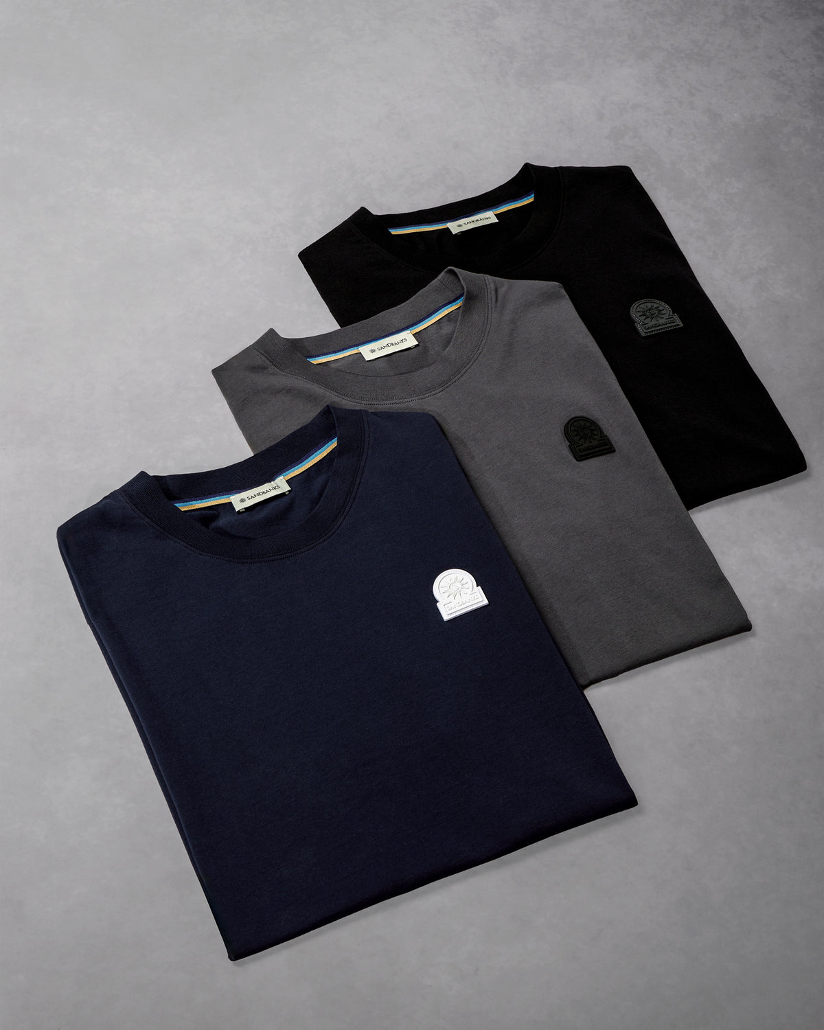 Three folded short-sleeve crewneck T-shirts arranged diagonally on a textured gray background. The shirts are navy blue, dark gray, and black, each featuring a small tonal chest patch and a thin multicolor stripe detail inside the collar.