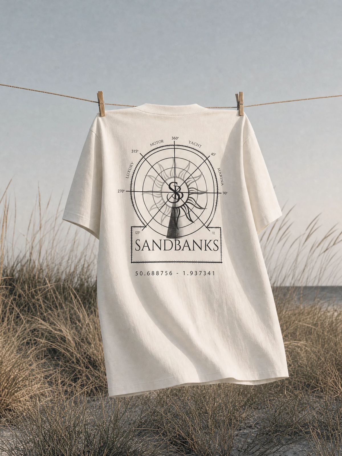 An off-white t-shirt hanging on a clothesline outdoors in a beach dune setting, featuring a black graphic print of a compass and the word 'SANDBANKS' on the back.