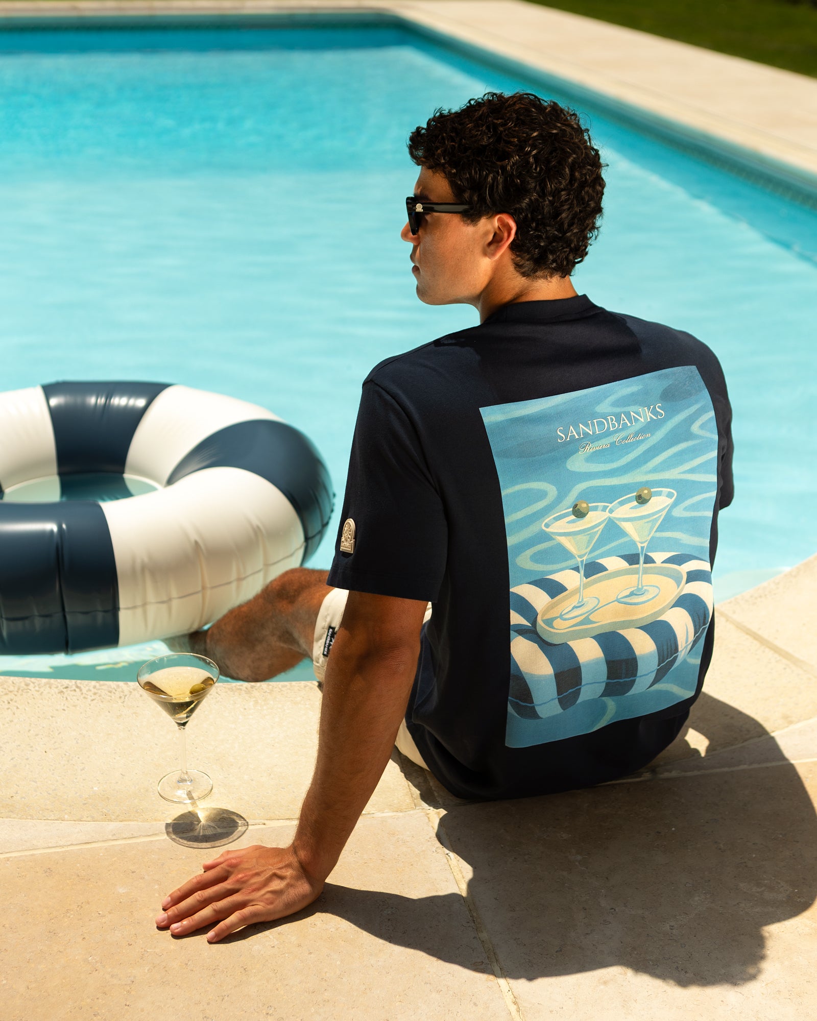 A man wearing a navy blue t-shirt with a large graphic print sits on the edge of a swimming pool. The back of the shirt features a light blue and white design with cocktail glasses 
