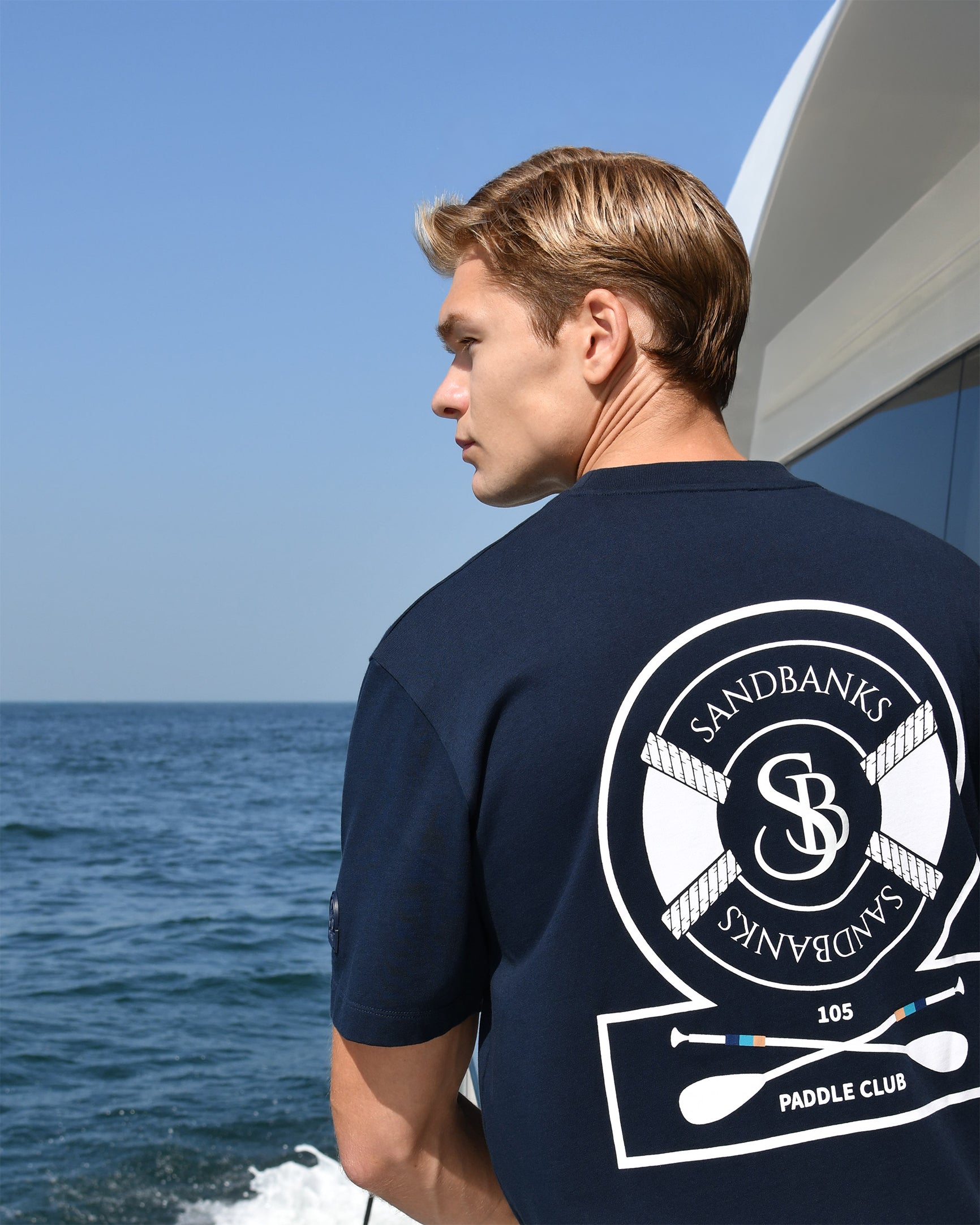 Man wearing a navy blue t-shirt with Sandbanks Paddle Club logo on a boat.