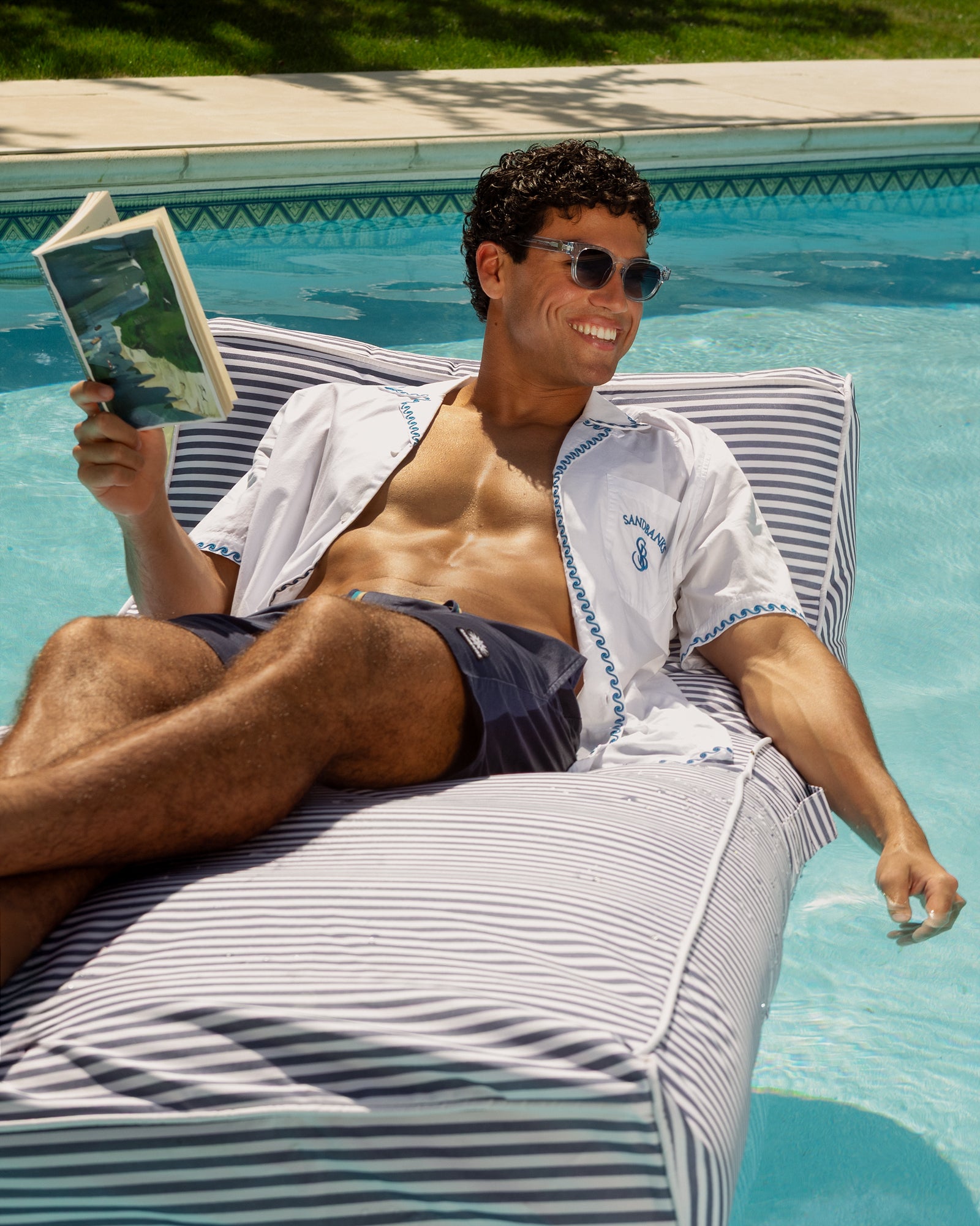 A smiling man wearing sunglasses and an open white short-sleeve shirt lounges on a striped pool float beside a swimming pool, holding an open book.