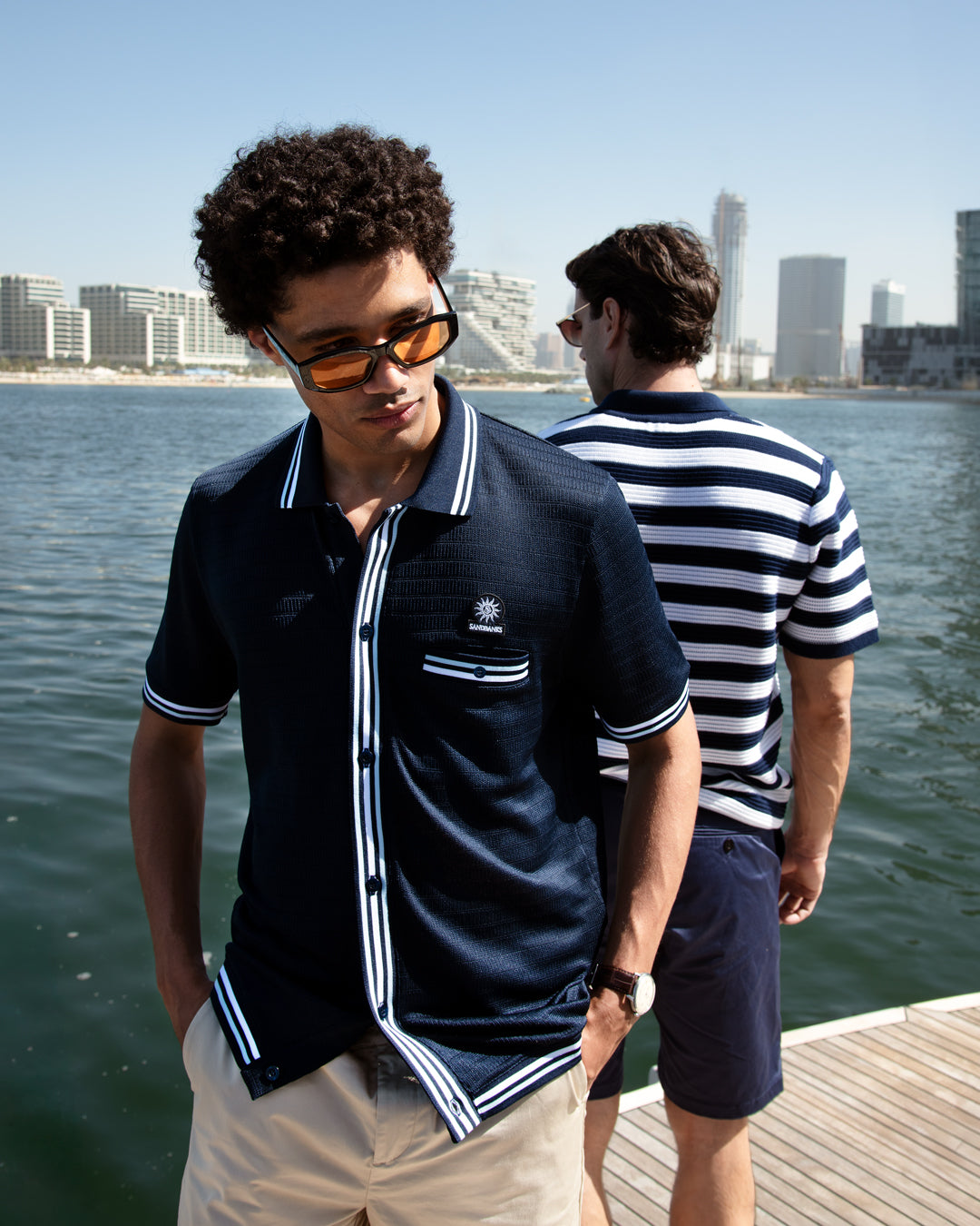Two men standing on a dock by water with city skyline in the background