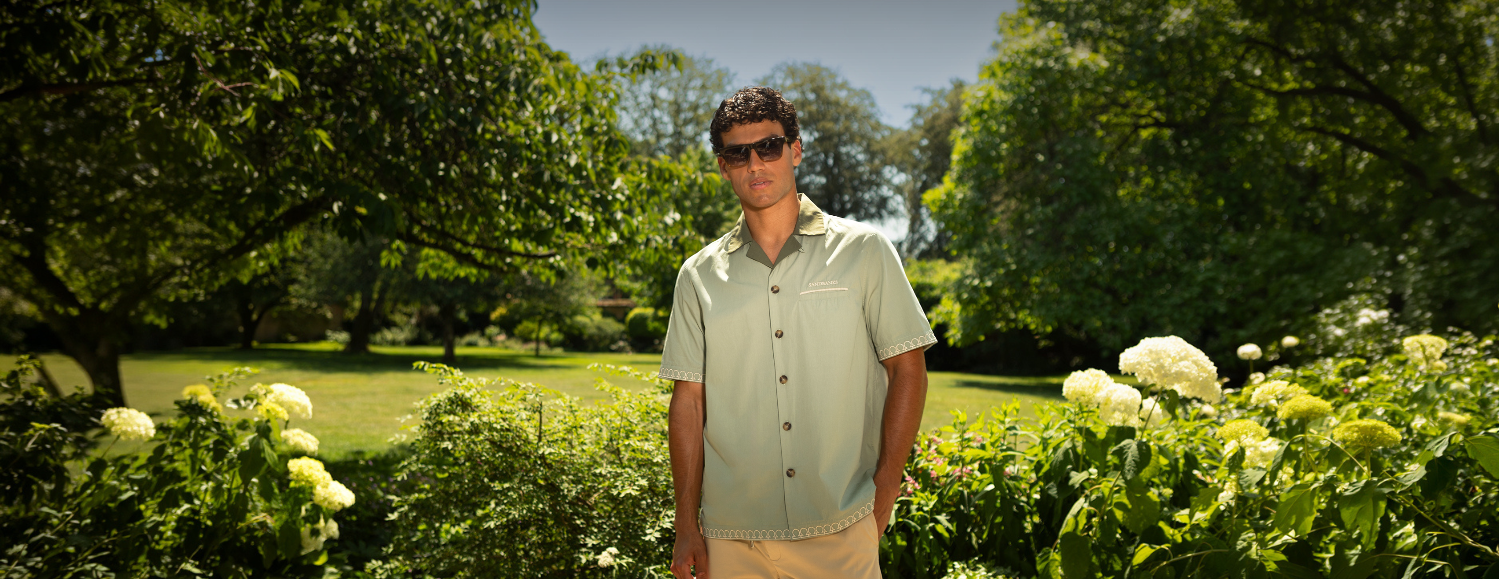 A man wearing a light sage green short-sleeve button-up shirt and beige pants stands in a lush green garden with white flowers and trees in the background.