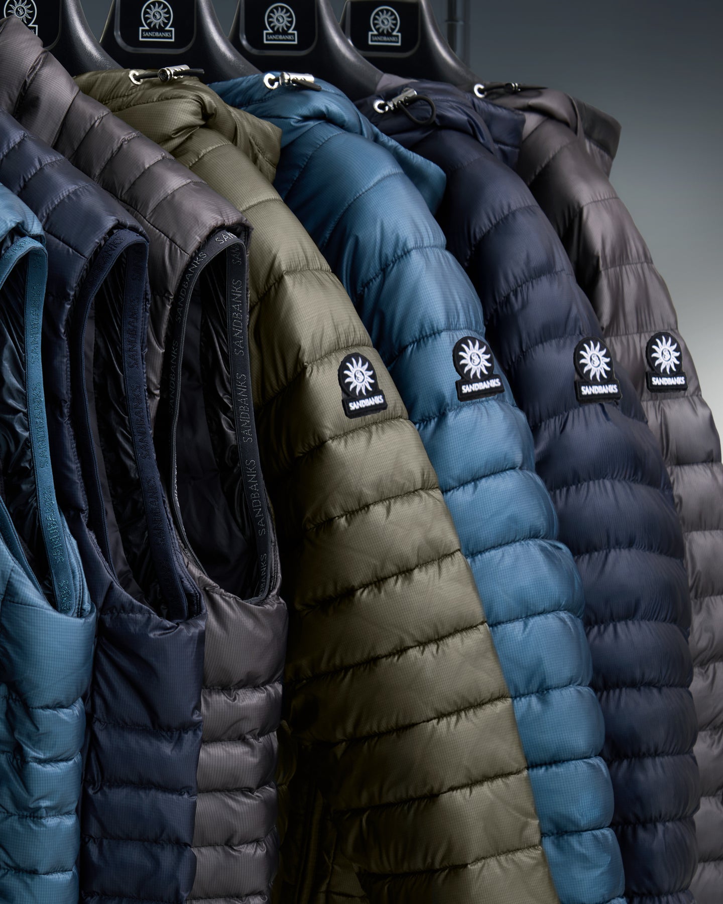 A close-up of several puffer jackets hanging on a rack, showing the brand patch. The jackets are in dark blue, olive green, slate blue, and dark gray.