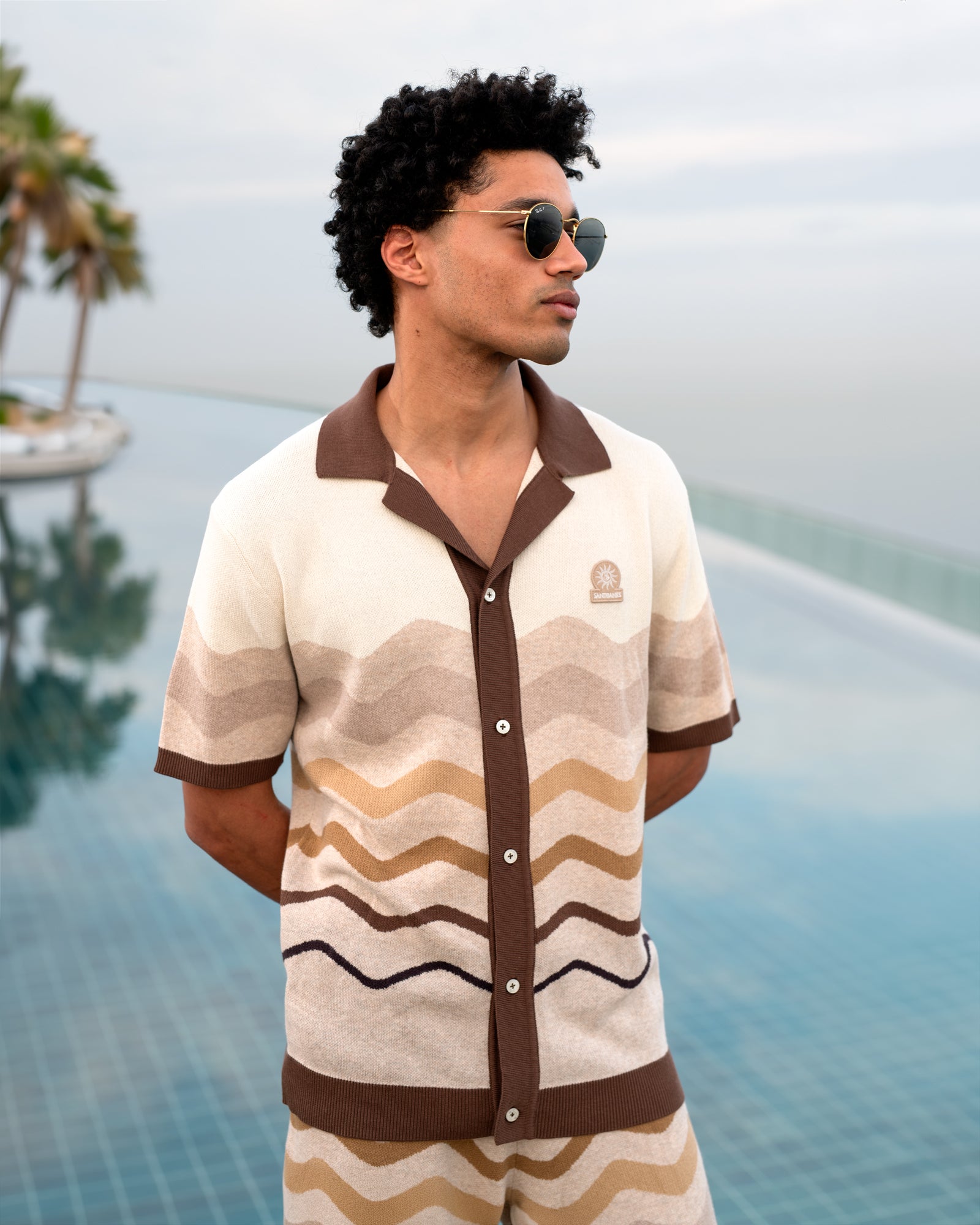 Man wearing a patterned shirt and sunglasses by a pool with palm trees in the background