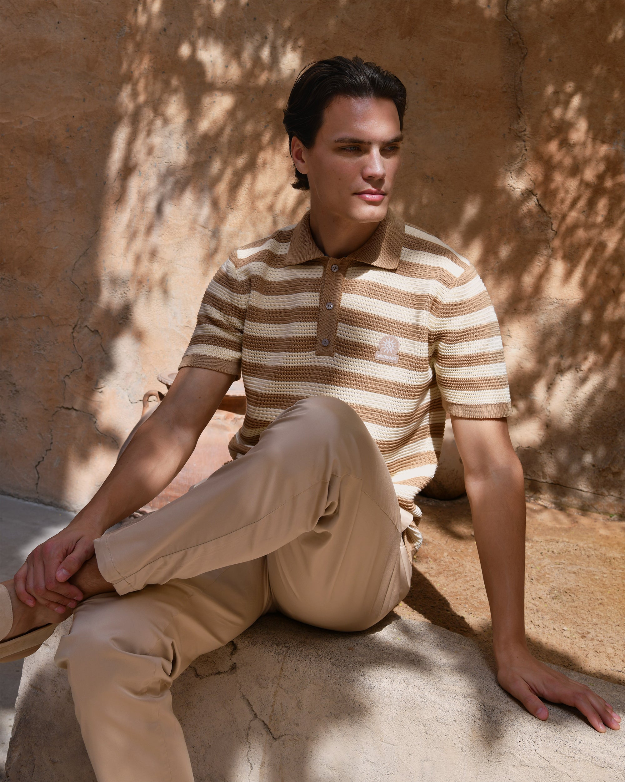 Man wearing a striped polo shirt and beige pants sitting against a textured wall.