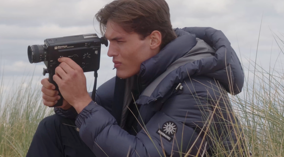 A man wearing a navy blue puffer jacket with a gray hood sits in tall grass outdoors, holding a vintage black film camera. The jacket has a visible circular logo patch on the sleeve that reads 'SOL'S'.
