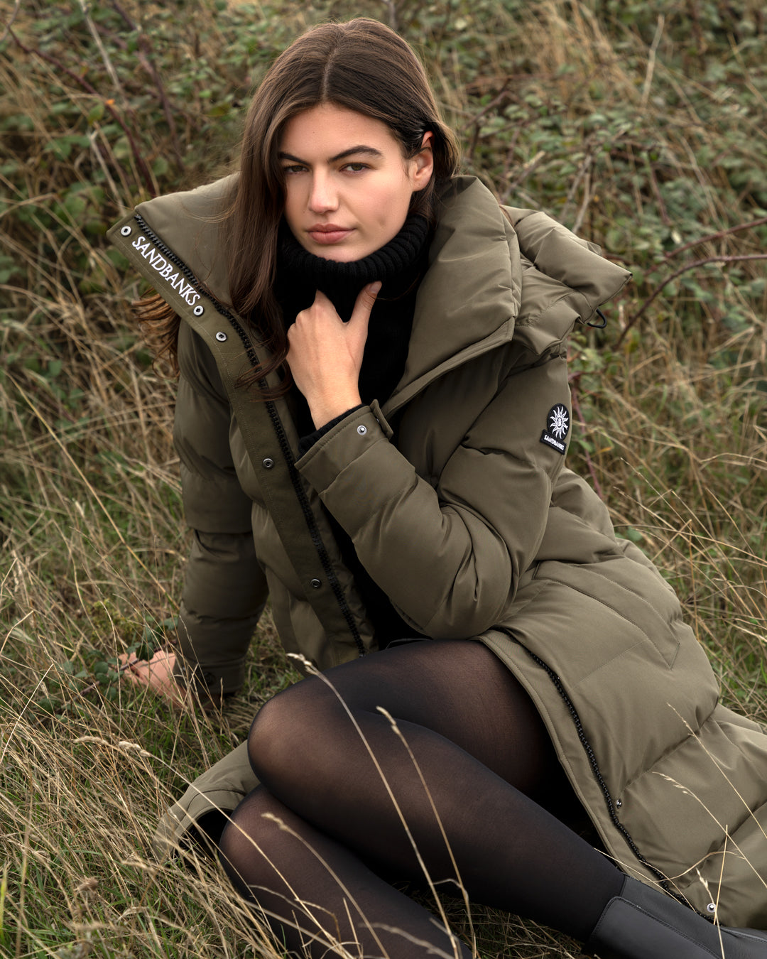 A woman wearing a long olive green puffer coat with a black turtleneck and black tights, kneeling in a grassy outdoor field. The coat features a high collar, front zipper and snap closure, and a visible 'SANDBRUNGS' brand patch on the shoulder strap.