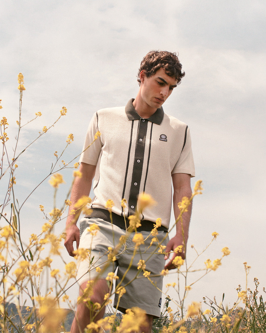 Man standing in a field of yellow flowers wearing a light-colored shirt with dark accents.